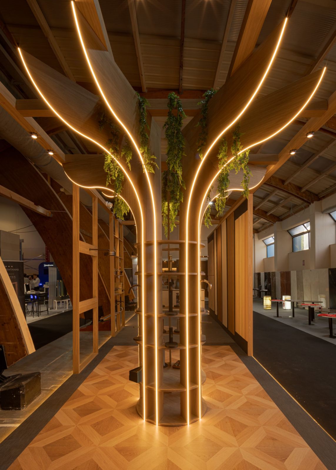 Vista lateral del stand El Santuario en Marbella Home Meeting 2026 con árbol-columna LED, pasillo entre arcos de madera y pavimento de marquetería, por Klic Arquitectos